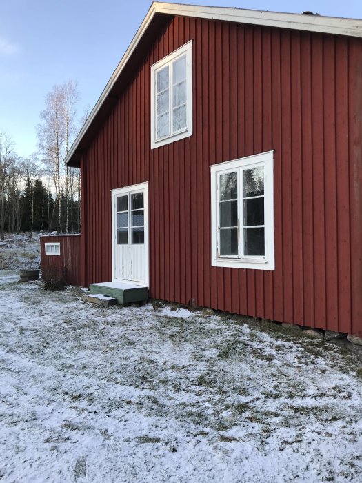 Röd timmerstuga från 1800-talet med vita knutar och fönster, snötäckt mark framför, klar för en tillbyggnad.