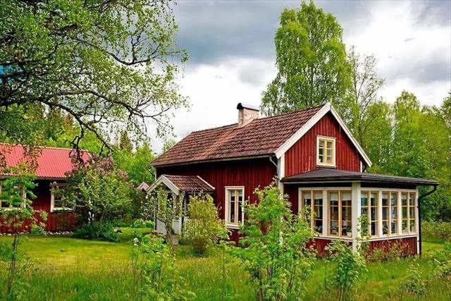 Rött traditionellt hus med vita knutar och veranda omgivet av grönska.