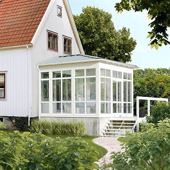 Vitt torp med modern glasveranda som påbyggnad, omgiven av grönska och trädgårdsväxter.