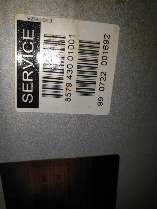 Etikett med serviceinformation och streckkoder på en apparat, suddig bakgrund.