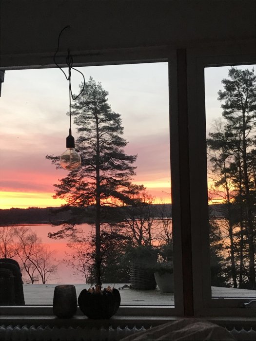 Utsikt från ett fönster i ett gammalt hus vid solnedgång, med silhuetten av träd och en sjö i bakgrunden.