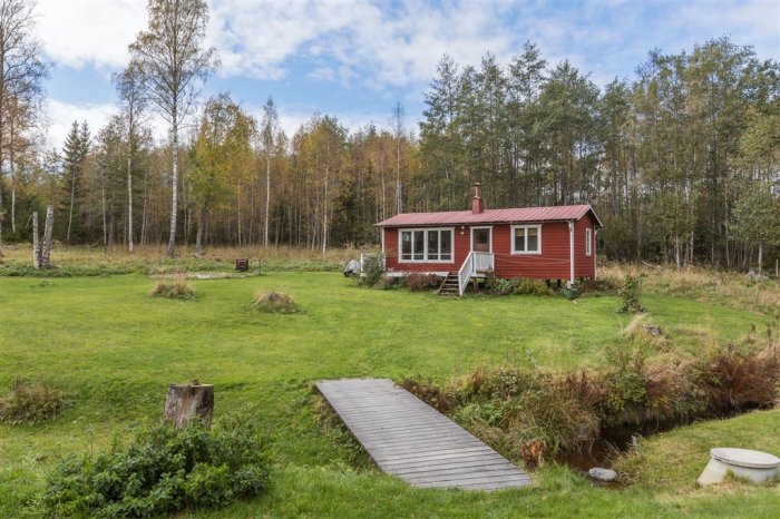 Rött gästhus med vita knutar omgivet av grönt gräs och skog, en liten bäck syns i förgrunden.