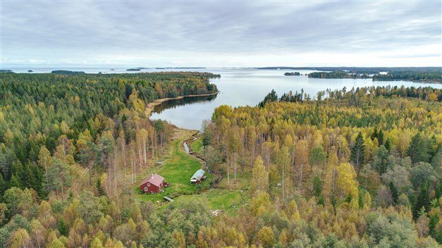 Flygbild över landskap med röd stuga, gästhus och bäck som leder ner till havet.