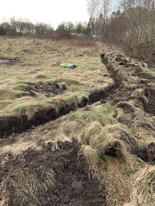 Nygrävd fiberkabeldike i gräsmark med synlig märkningsspray och grävskaft i bakgrunden.