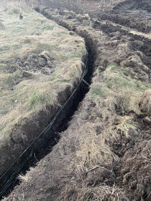 En djup grävd grävning i en trädgård som exponerar en fiberoptisk kabel, omgiven av gräs och jord.