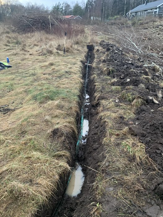 Grävd grön fiberkabel synlig i en lång grävning över en gräsmark med vattenansamling.