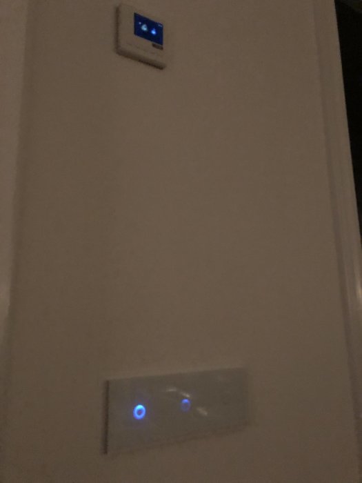 En termostat och strömbrytare med upplysta knappar på en vägg i svagt ljus.