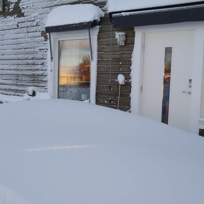 En täckt snöyta framför hus med snöbelagd fasad, dörr och fönster som speglar solnedgången.