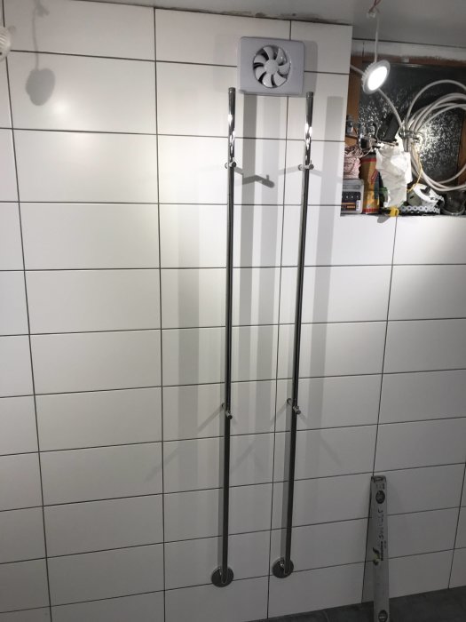 Nylagda vita kakelplattor på vägg med monterad ventilation och två vertikala metalldetaljer.