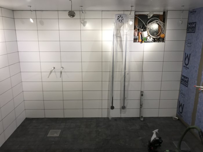 Renoverat badrum med vita kakelväggar och mörkgrått golv, installation ej helt färdig, byggmaterial synligt.