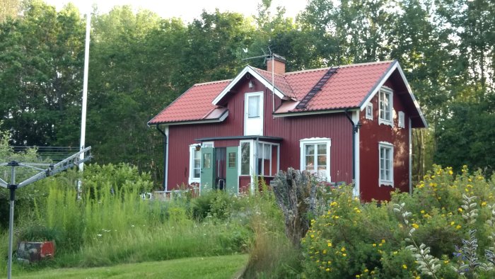 Röd trävilla med liten utbyggnad framför huset, omgiven av grönska och blommor.