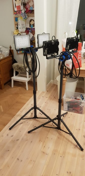 Två LED-bygglampor monterade på ett svart stativ med slingrade kablar, inomhus på ett trägolv.