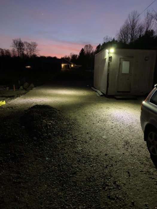 Arbetsbod upplyst av nyinstallerad arbetsbelysning i skymningen med ett grusområde i förgrunden.