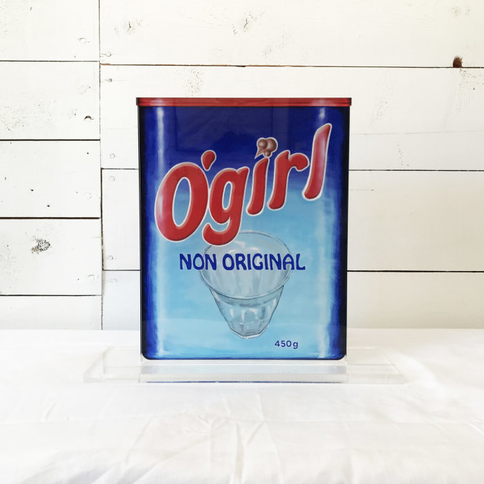 Blå förpackning med texten "O'girl NON ORIGINAL" och bild av glas, framför vit träpanel.