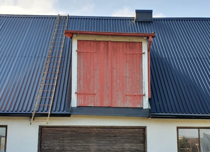 Rödmålad ladugårdsdörr på övervåning med stege lutad mot blåtakad byggnad.