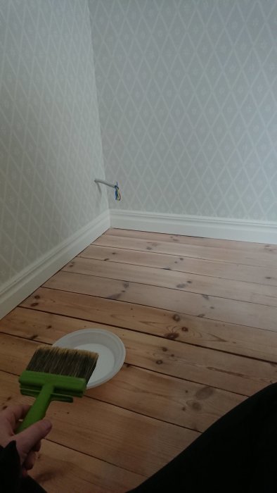 En hand håller en pensel över en tallriksassiett bredvid nylagt trägolv och nytapetserade väggar i ett rum under renovering.