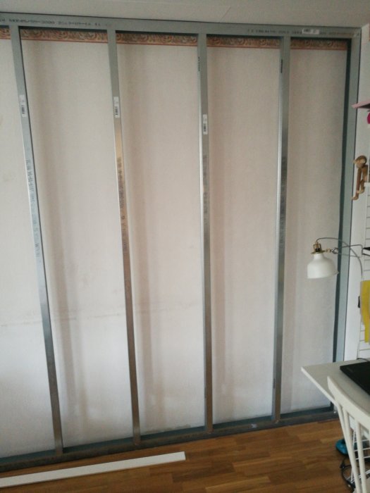Påbörjat bygge av vägg med smala, stående metallreglar uppsatta mot en befintlig vägg i ett rum.