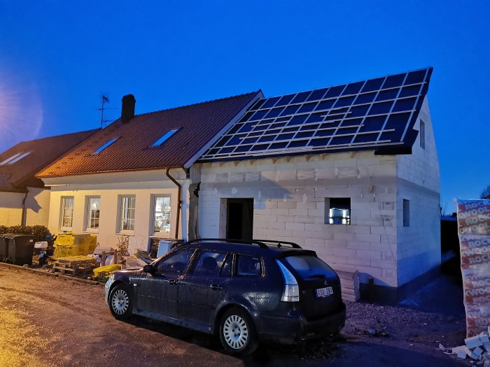 Hus under renovering med solpaneler på taket vid skymningen, bil parkerad framför.