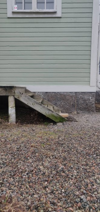 Sliten trätrappa med vit stolpe framför grön husvägg och grusad mark.