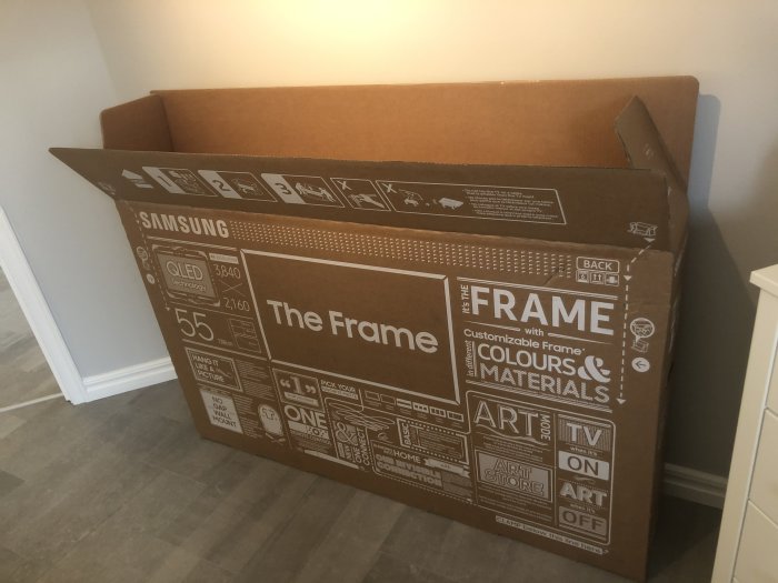 En stor kartong för en Samsung The Frame TV står lutad mot en vägg i ett hem.