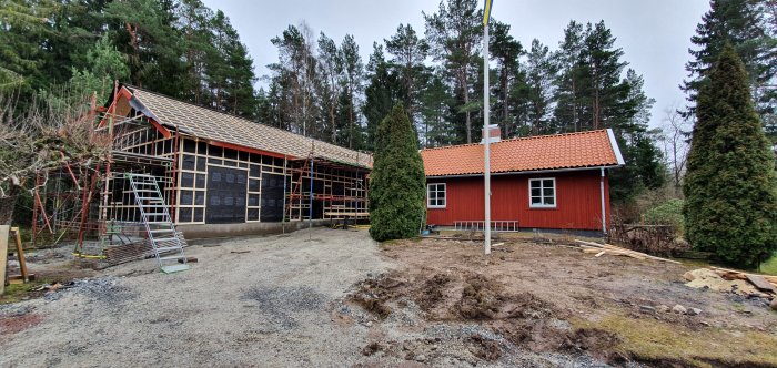 Rött hus med nytt tak under konstruktion, omgiven av träd och ställning, under molnig himmel.