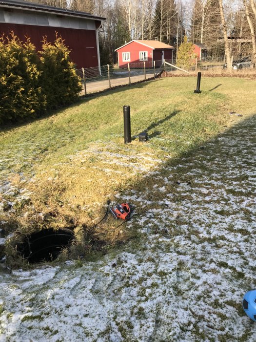 Bakgård med en öppen pumpbrunn, två vertikala rör och snötäckt gräs med hus i bakgrunden.