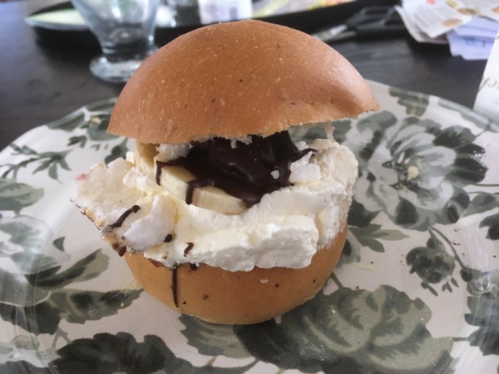 En semla med banan, grädde och chokladsås på ett mönstrat fat.