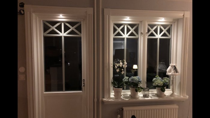 Två vitmålade fönster med krysstag och en Hide-a-lite LED Dot VP spot ovanför, inomhusdekoration med blommor och en liten lampa på fönsterbrädan.