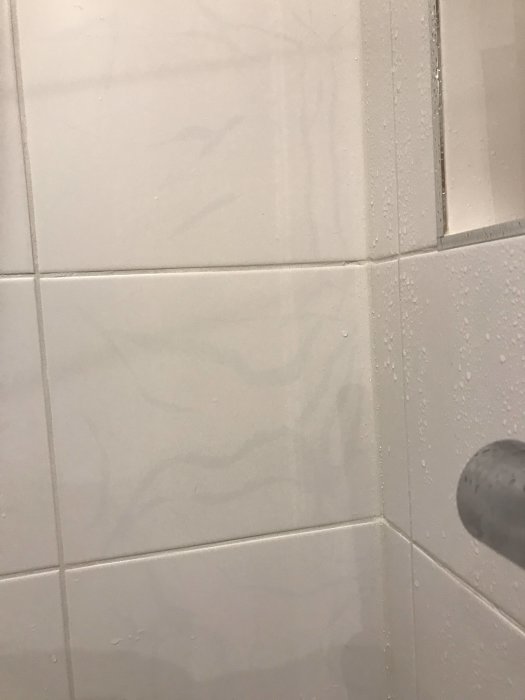 Vit kaklad vägg i dusch med vattendroppar och en del av duschhuvudet synligt.