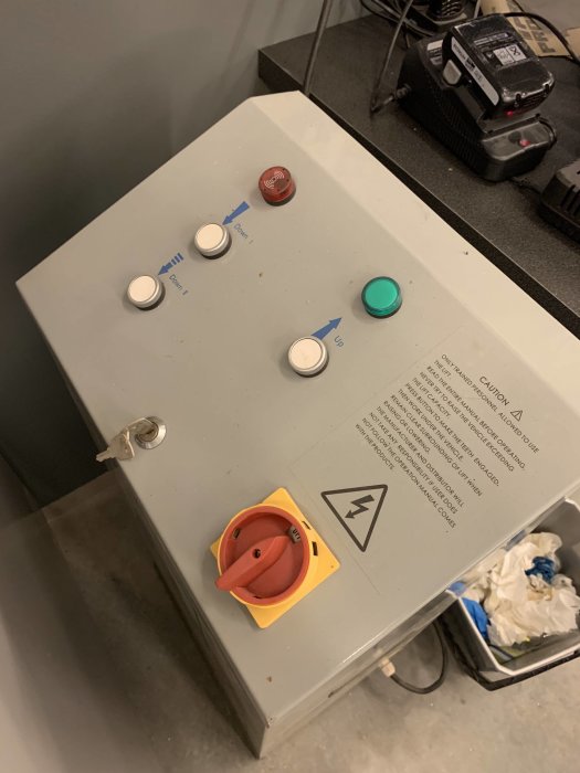 Kontrollpanel för lyft med knappar märkta "Upp/Ner", nödstopp och strömbrytare, i en verkstadsmiljö.