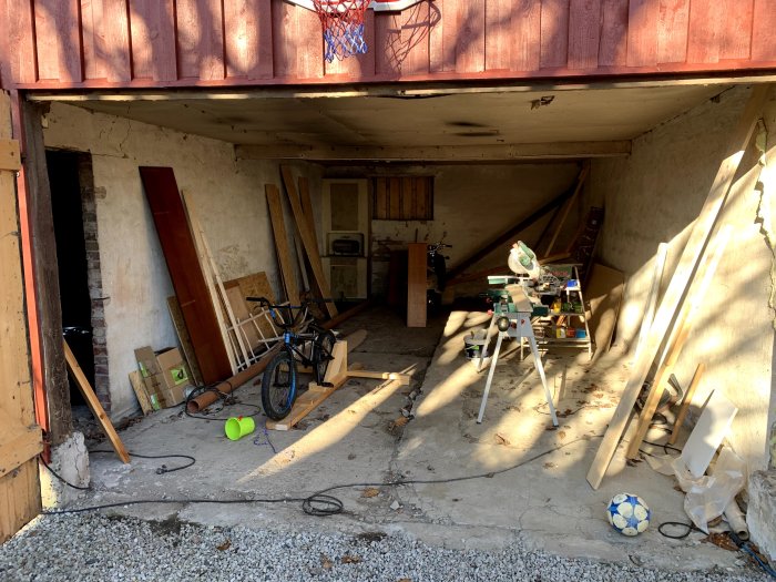 En verkstad i ett laduhus med ojämnt betonggolv i två nivåer, verktyg, brädor och en cykel syns.