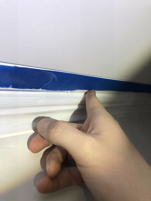 En person drar bort blå maskeringstejp från en vitmålad fog mellan taklist och innertak.
