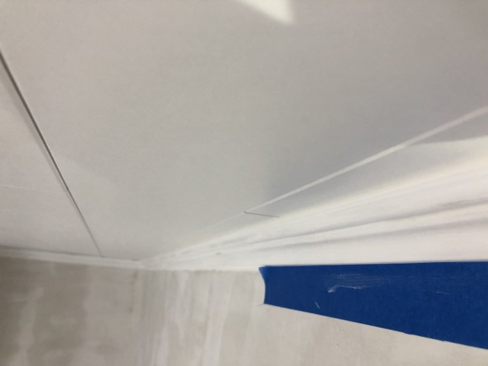 Hörnet av ett innertak med en vit folierad yta anslutande mot en taklist, skyddad med blå maskeringstejp inför fogning.