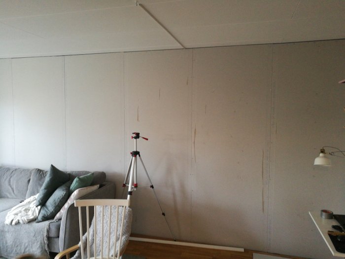 Oisolerad vägg delvis täckt med gipsplattor, synliga skruvlinjer och en tripod framför.