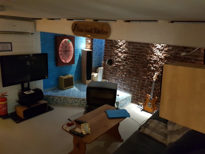 Inredd källare med belysning på tegelvägg, TV, gitarr, kamin och stort väggur.