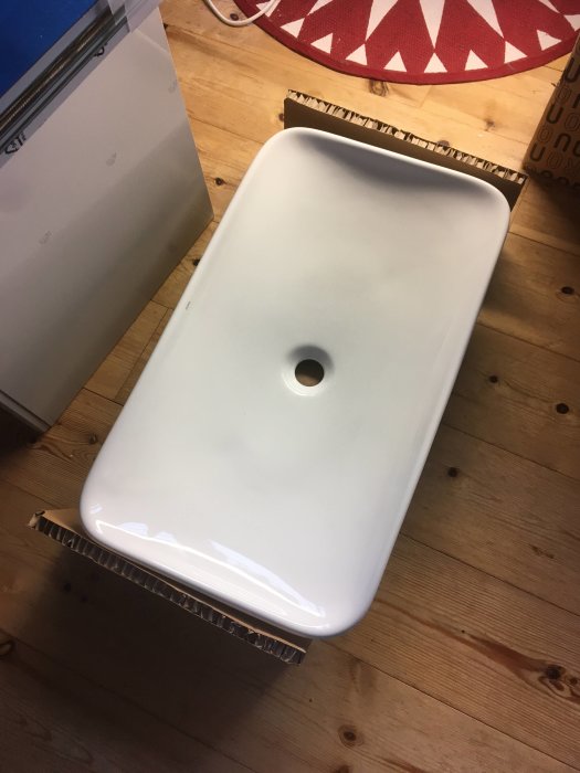Nytt vitt handfat som fortfarande ligger i kartongen på ett trägolv, upplagt hemma före installation.