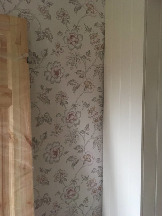 Del av en vägg med ljus tapet med lantligt, romantiskt blommönster, intill en vit dörrkarm.