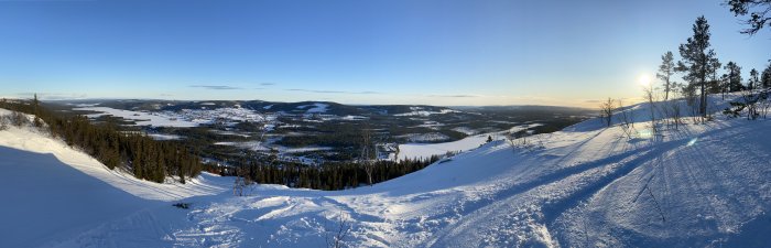 Panoramavy över ett snöklätt landskap med träd, berg och en klarblå himmel vid solnedgång.