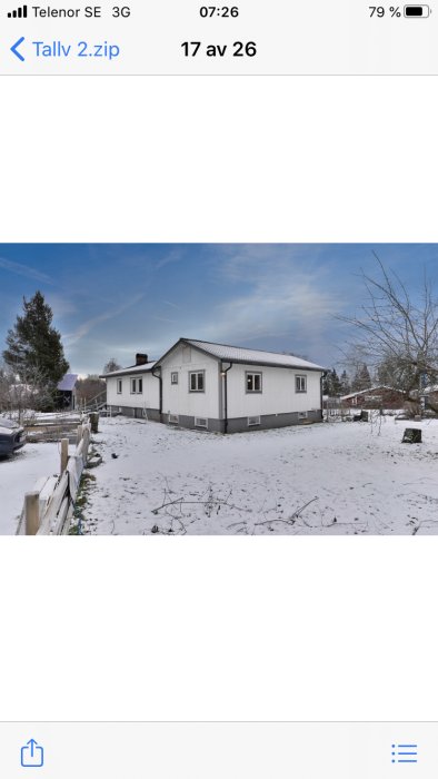 Vit enplansvilla med svart tak täckt av snö, omgivet av vinterträdgård och staket.