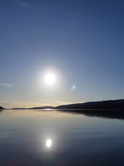 Solen speglar sig i en lugn sjö med kullar i bakgrunden under en klarblå himmel.