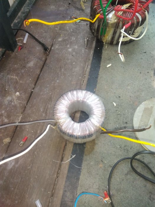 Toroidal transformer next to a large ring transformer with wires on a wooden floor.