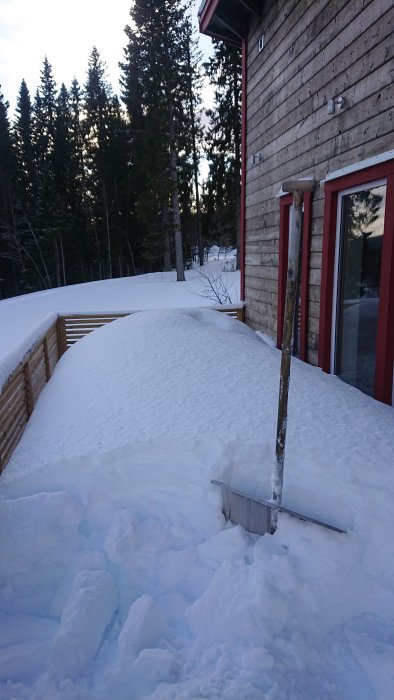 Altan delvis täckt av snö med skottade högar intill trävägg och räcke i snöigt landskap.