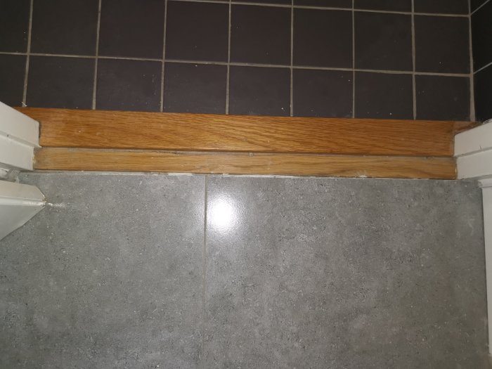 Tröskel i ek mellan grått klinkergolv och ljusgrått golv, med synlig glipa vid dörrkarmen.