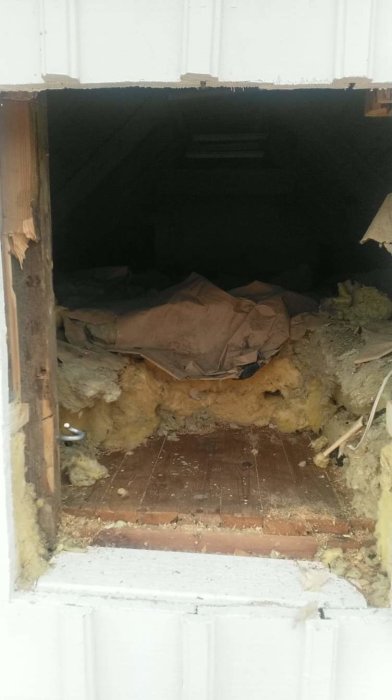 Utsikt genom en vindslucka med gamla isoleringsmaterial och skräp på en vind.