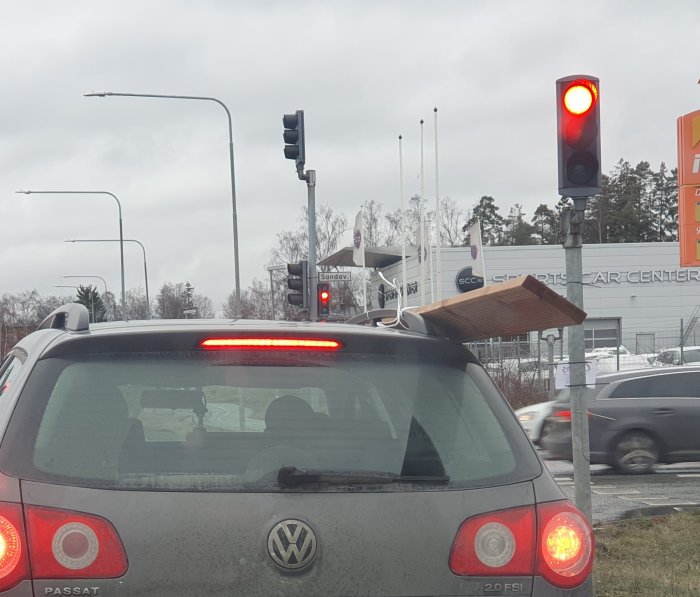 Bil vid trafikljus med långt träbräde som sticker ut genom bakrutan.