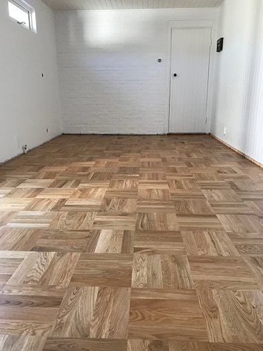 Ett tomt rum med rutformat parkettgolv från 1970-talet och vitmålade väggar och tak.