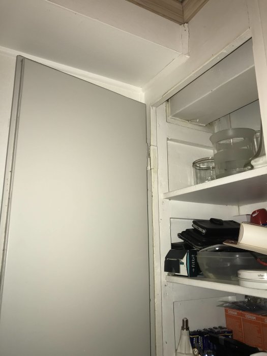 Grå låda som döljer ventilationskanalen i hörnet av ett kök med synliga hyllor och köksartiklar.