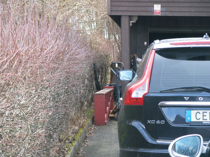 Garage med öppen dörr och bil framför, i en trång uppfart bredvid en häck.