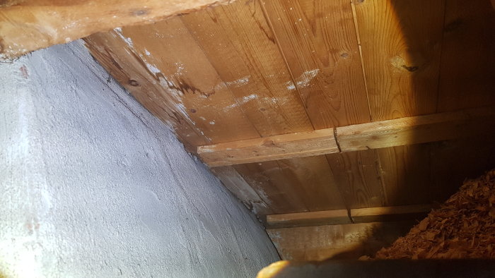 Träkonstruktion och spånskikt på ett kallvinds golv med fuktfläckar nära en skorsten.