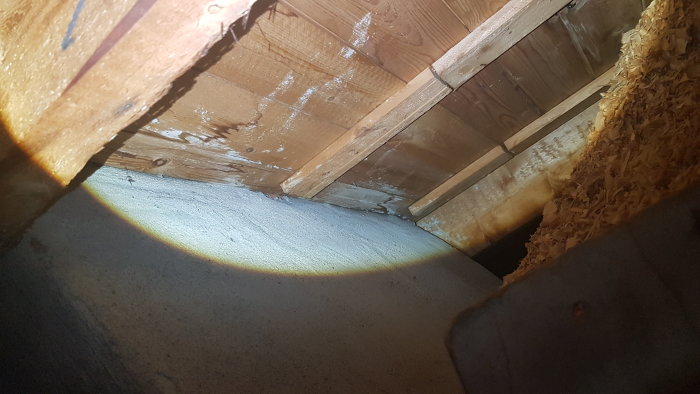 Kallvindsutrymme i hus byggt 1954 med synliga träbjälkar, sågspån på golvet och potentiella fuktfläckar vid skorsten.